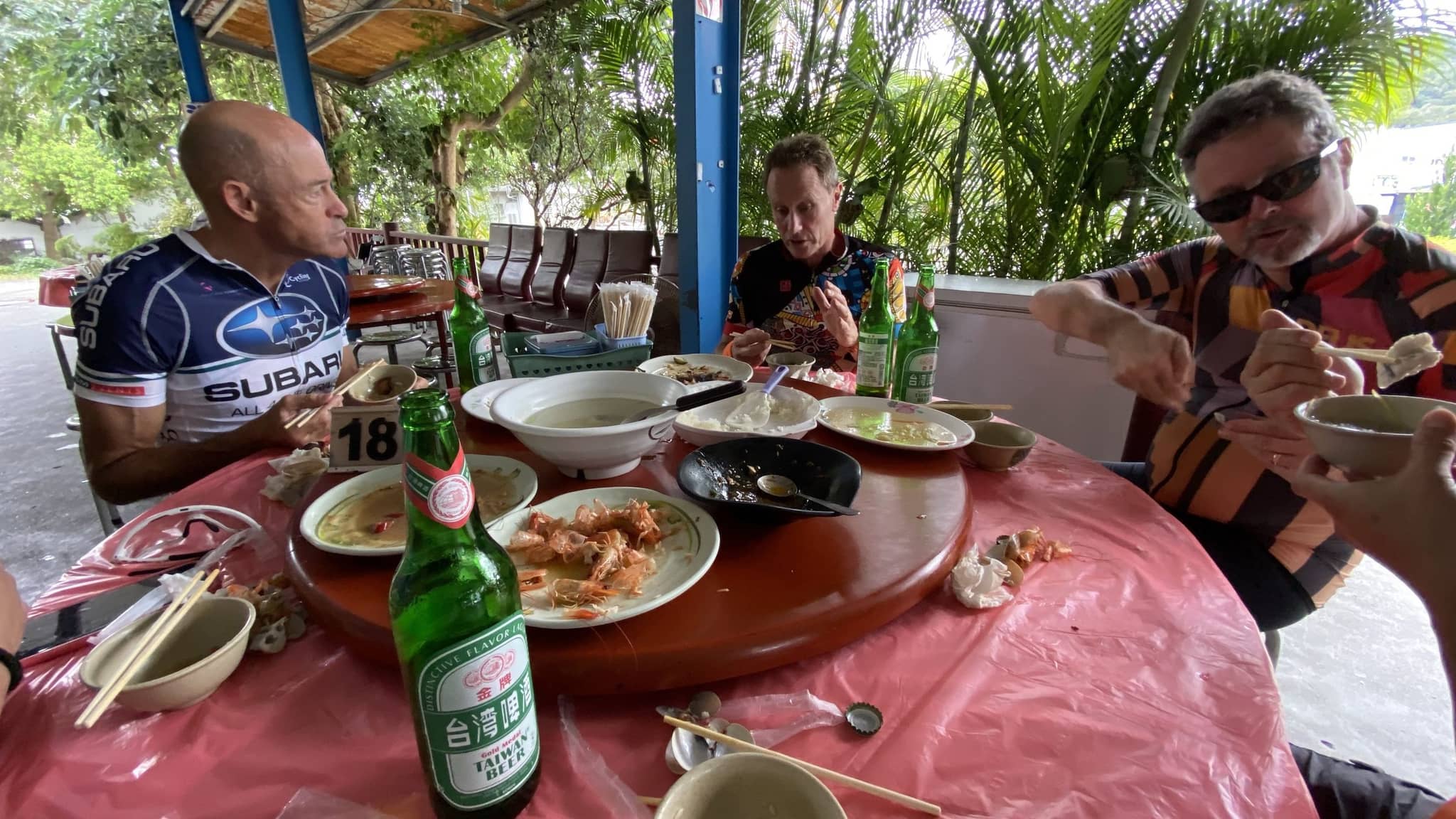 Three cyclists in jerseys eating dumplings and noodle soup at an outdoor restaurant table, bottles of Taiwan Beer on a red round dining table with palm trees behind