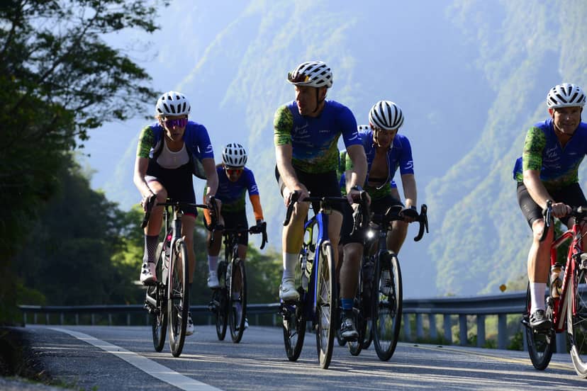 Group of cyclists riding together across a bridge with green mountains rising in the background