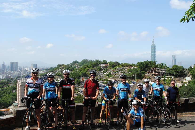 Cycling tour group posed on a hilltop above Taipei with the Fu De Keng cemetery in the foreground and Taipei 101 rising from the city skyline in the distance
