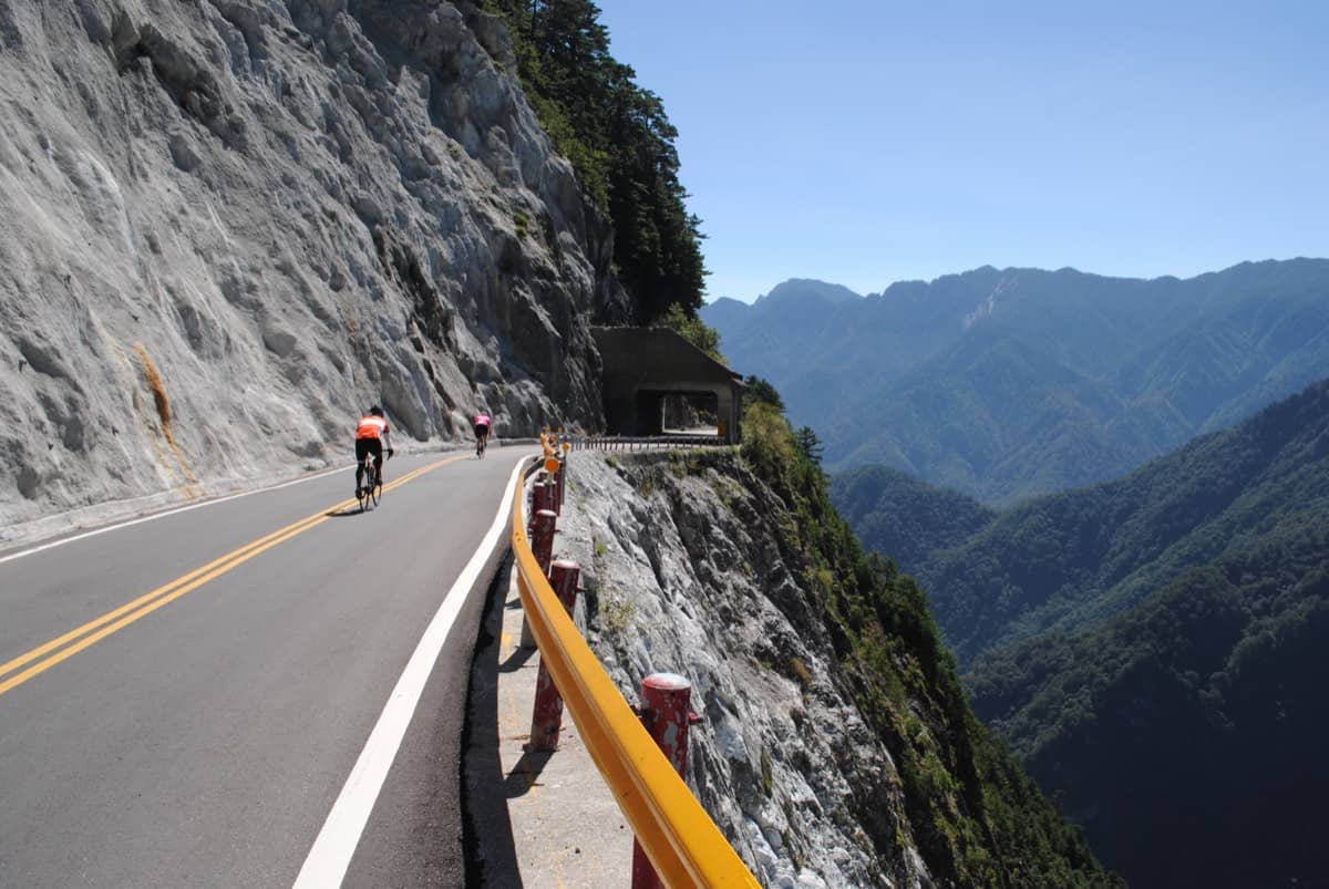 Two cyclists on a mountain road cut into a cliff face in Taiwan, deep blue-sky mountain views stretching into the distance