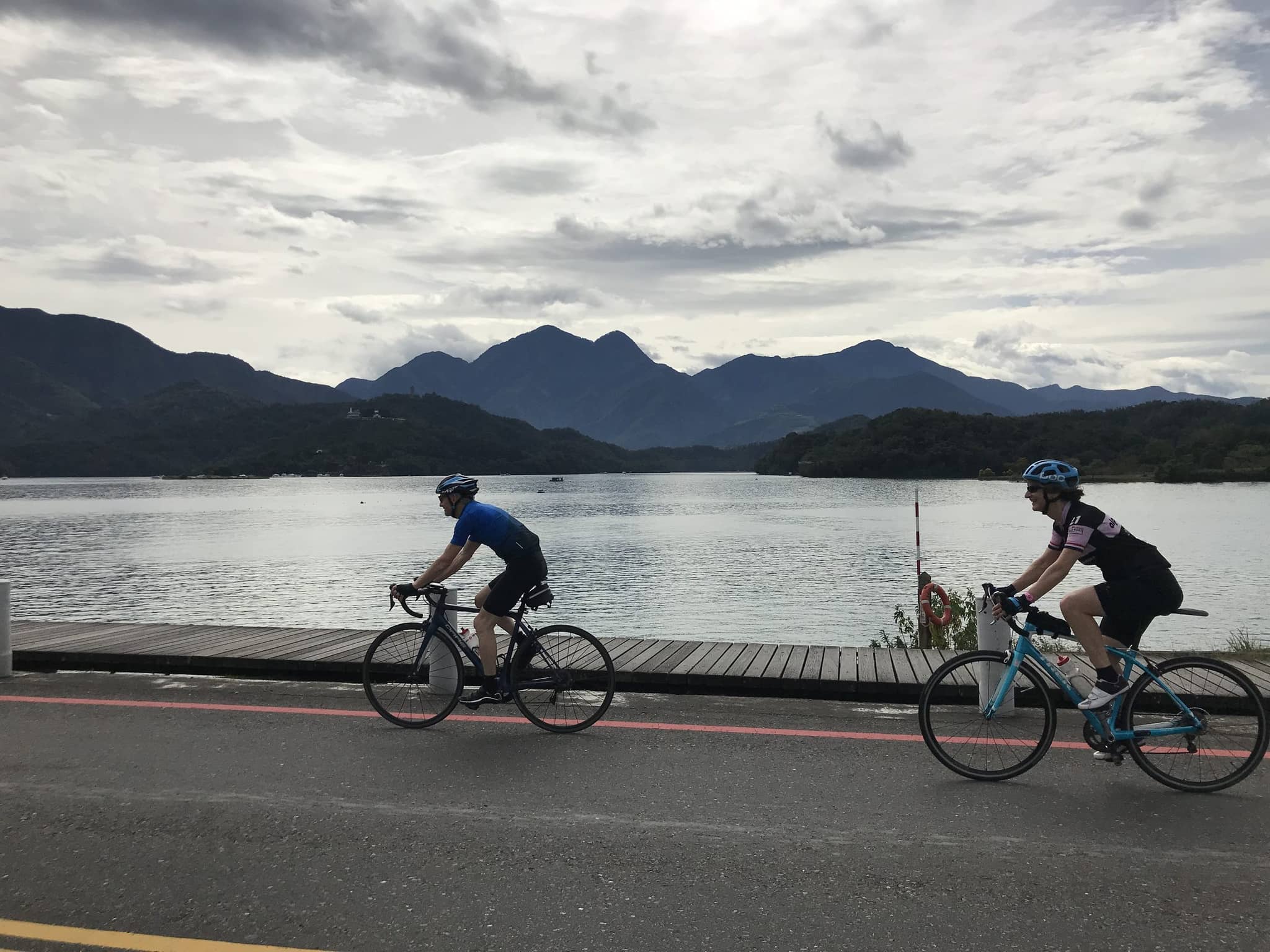 Two cyclists riding past Sun Moon Lake on the lakeside cycle path with the central mountain range and dramatic cloudy sky in the background