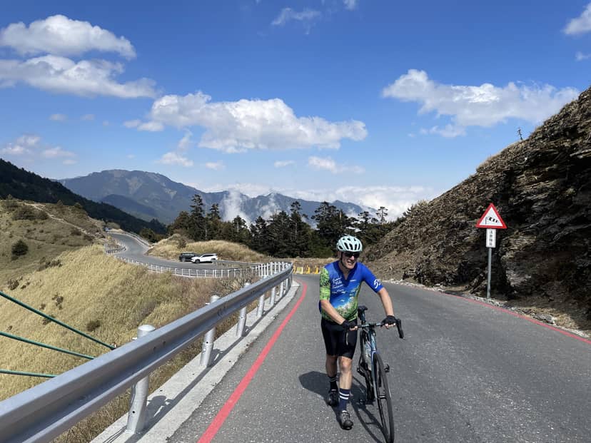 Cyclist smiling on a long climb near the summit of Hehuanshan on the Taiwan KOM route, distant peaks and blue sky behind