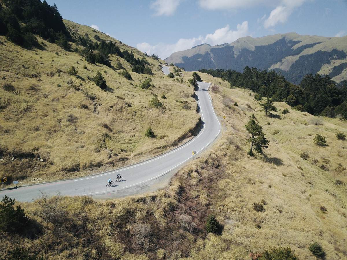 Aerial view of two cyclists riding a winding mountain pass road through golden alpine grassland in central Taiwan, with forested peaks in the distance