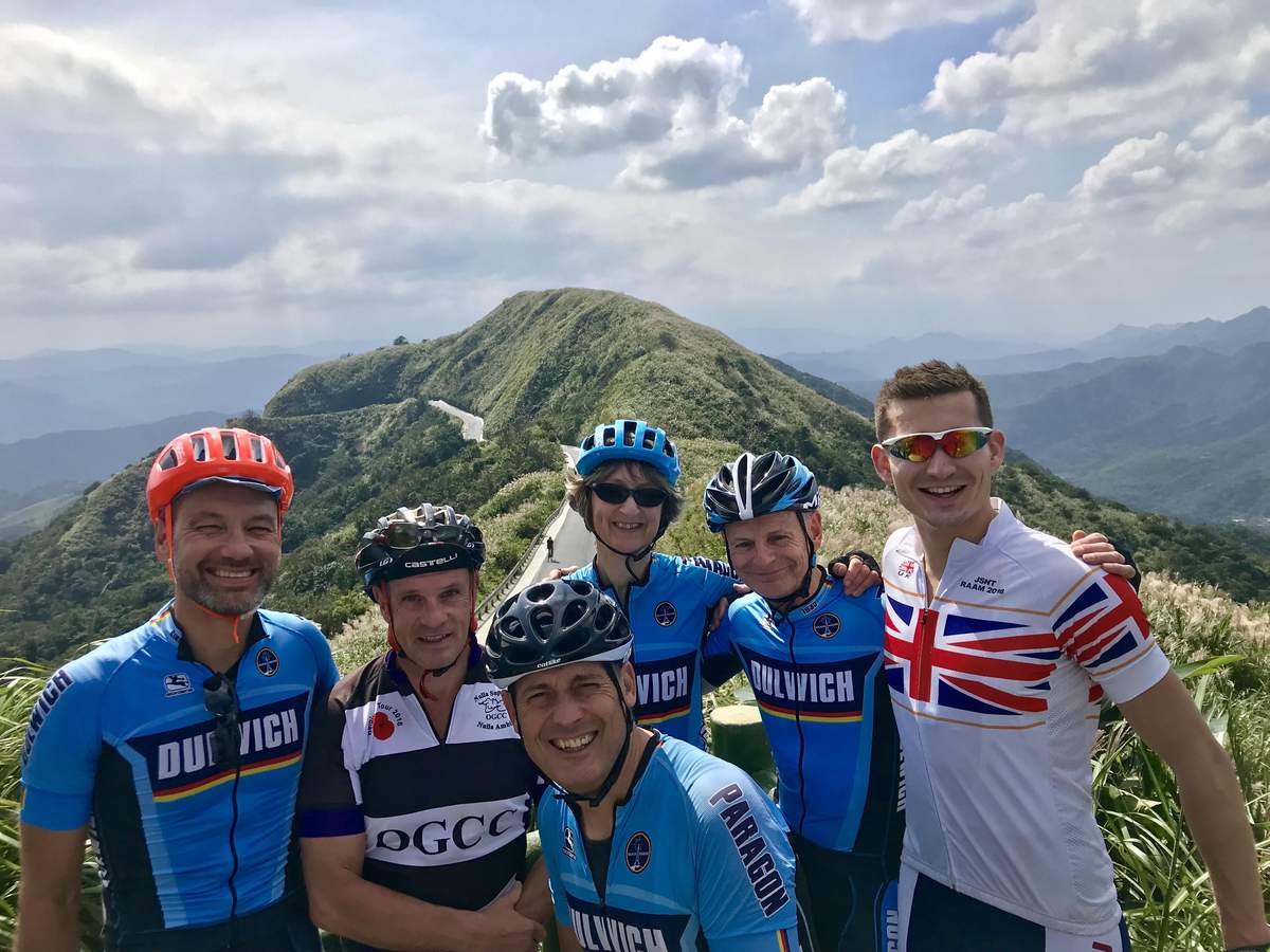 Group of cyclists posing for a summit photo on a Taiwan mountain top with green peaks behind