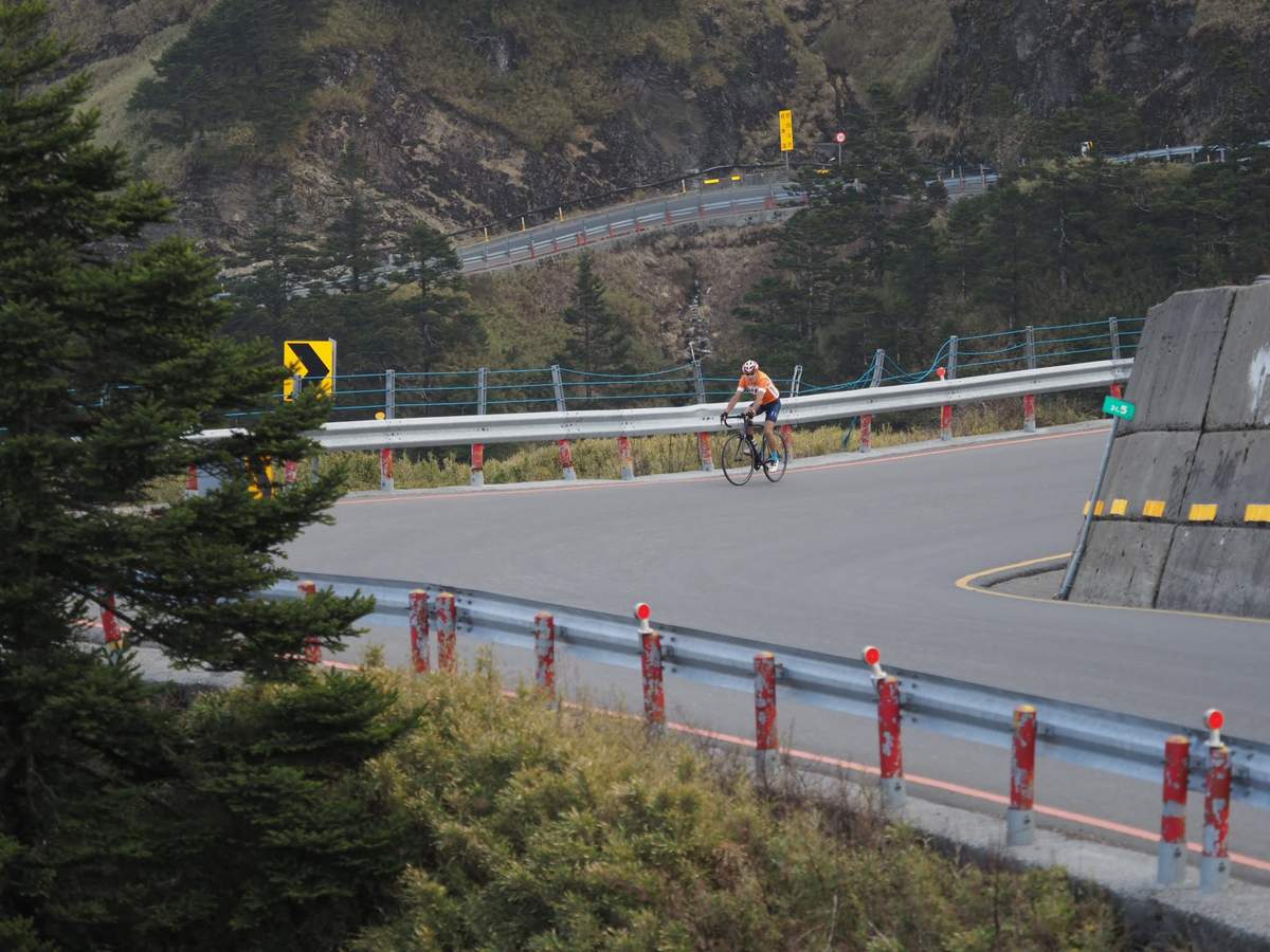 Cyclist on a switchback section of the upper Taiwan KOM route with chevron warning signs and concrete barriers, alpine terrain behind