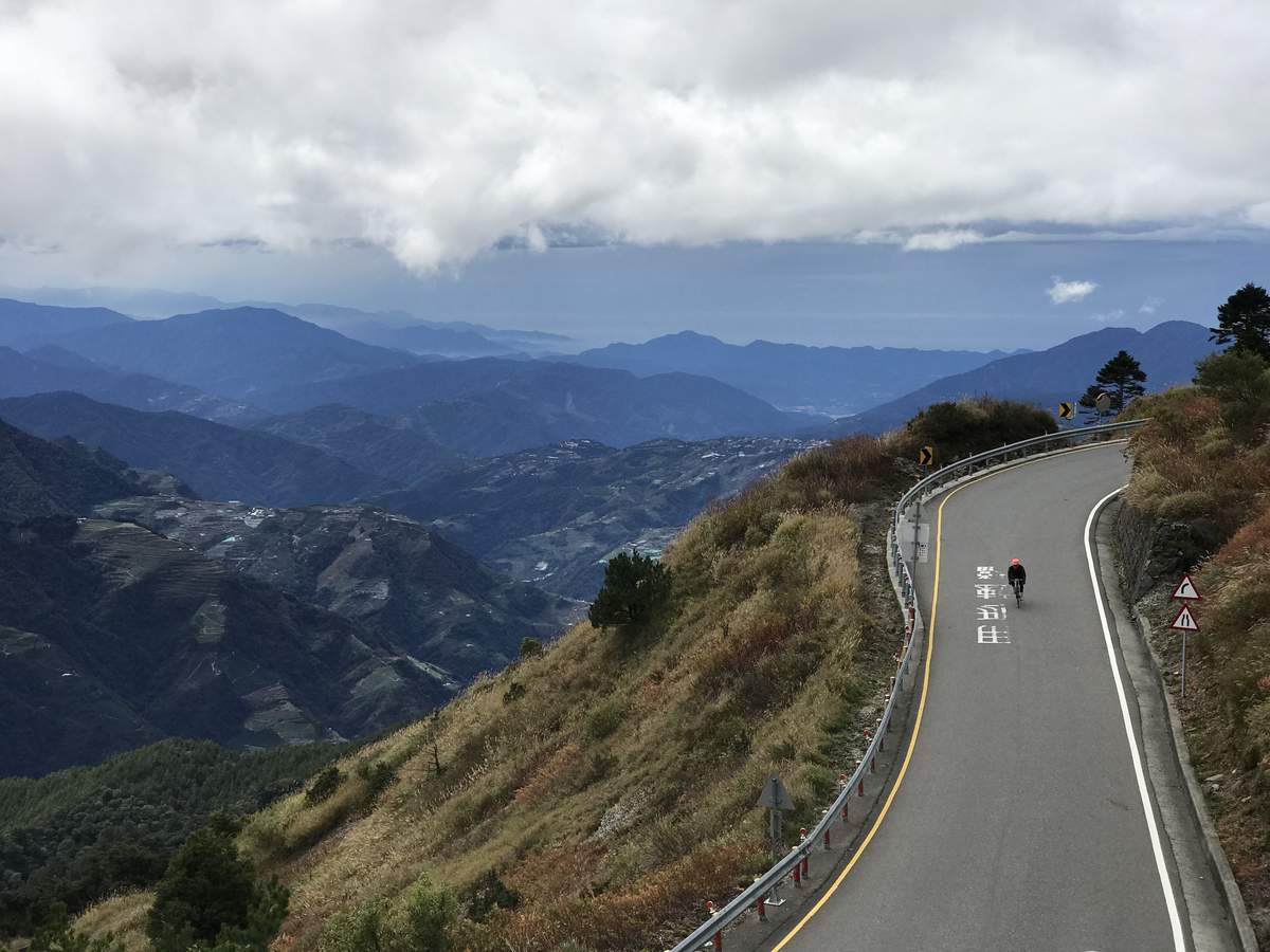 Cyclist descending a mountain switchback road with layered Central Range peaks dropping into cloud below