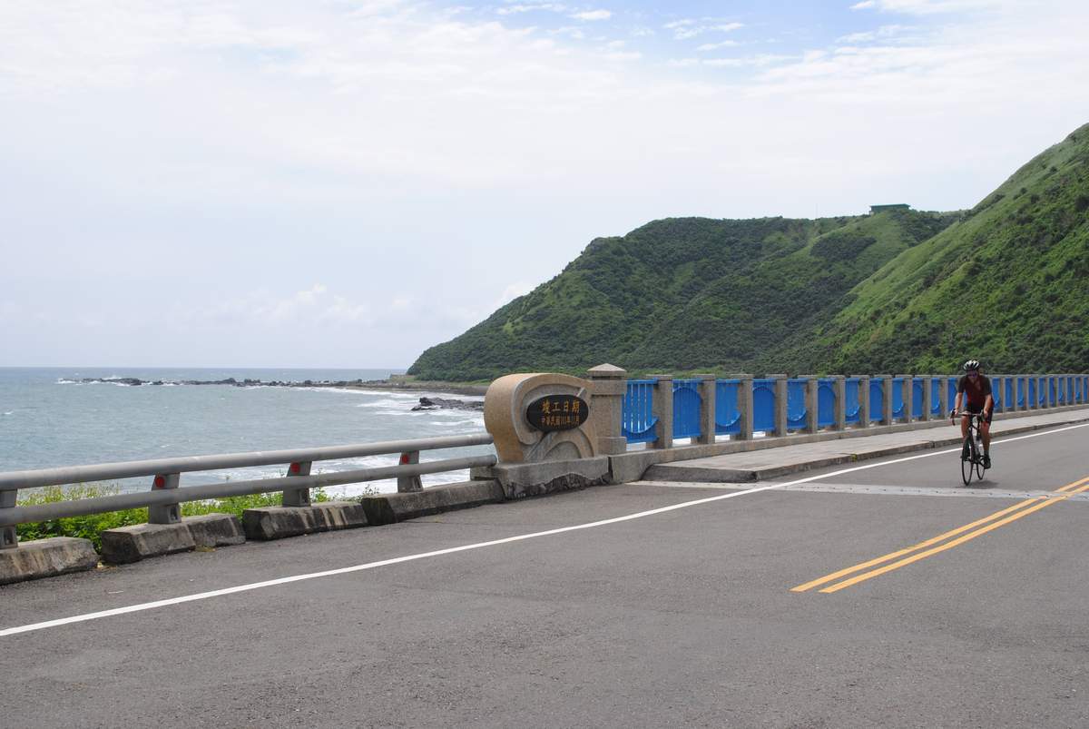 Cyclist on a coastal bridge with blue railings along the Pacific coast road south of Keelung, with rocky shoreline, calm sea, and green coastal hills in the background