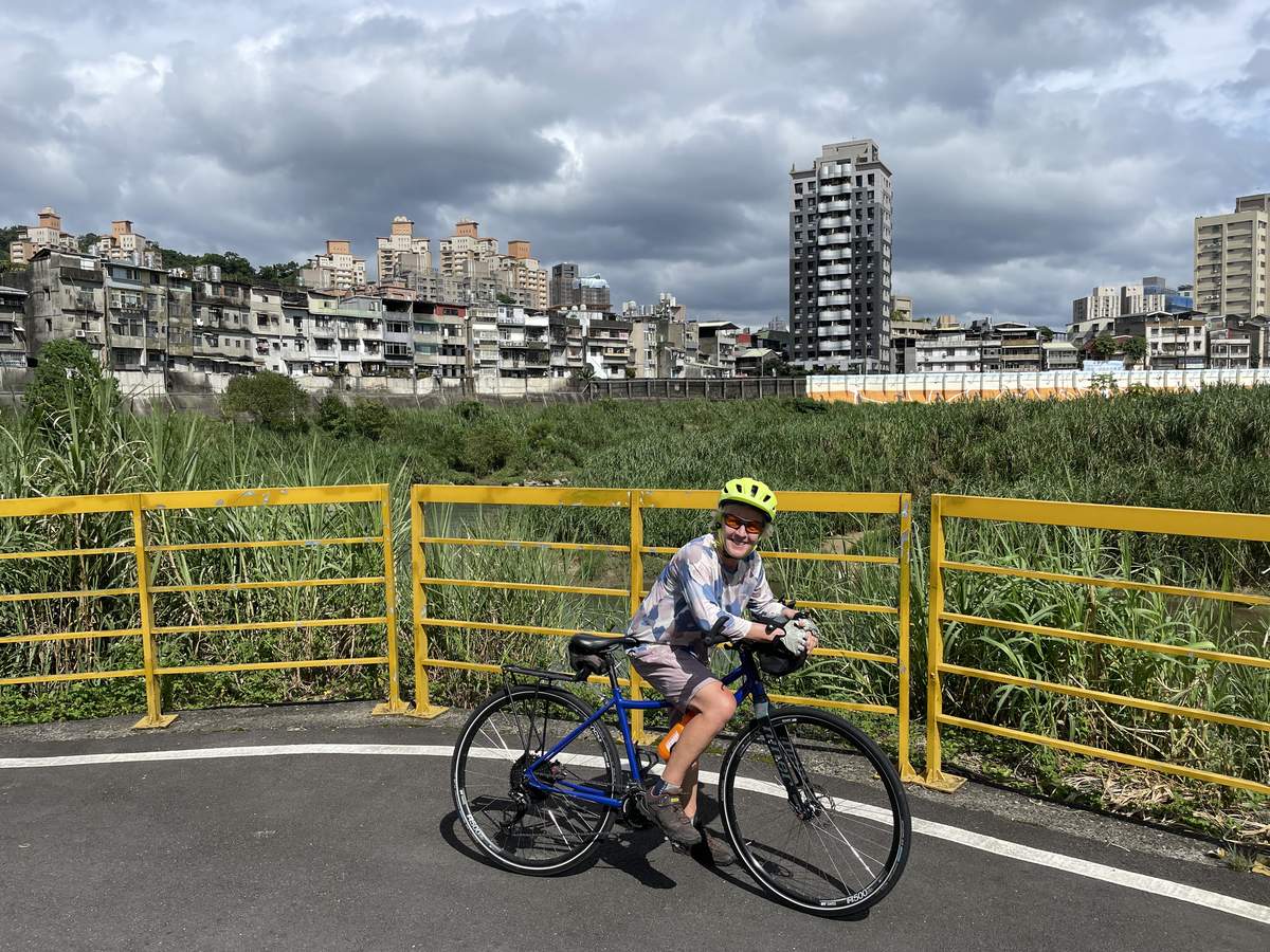 Solo cyclist on a Taipei riverside cycle path beside yellow railings, with reed beds in the foreground and apartment blocks under cloudy sky in the background