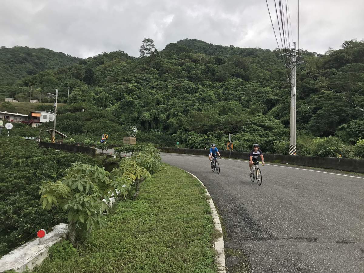 Two cyclists riding a quiet forested mountain road near Taipei under overcast sky, with lush green hillsides and small village houses in the background