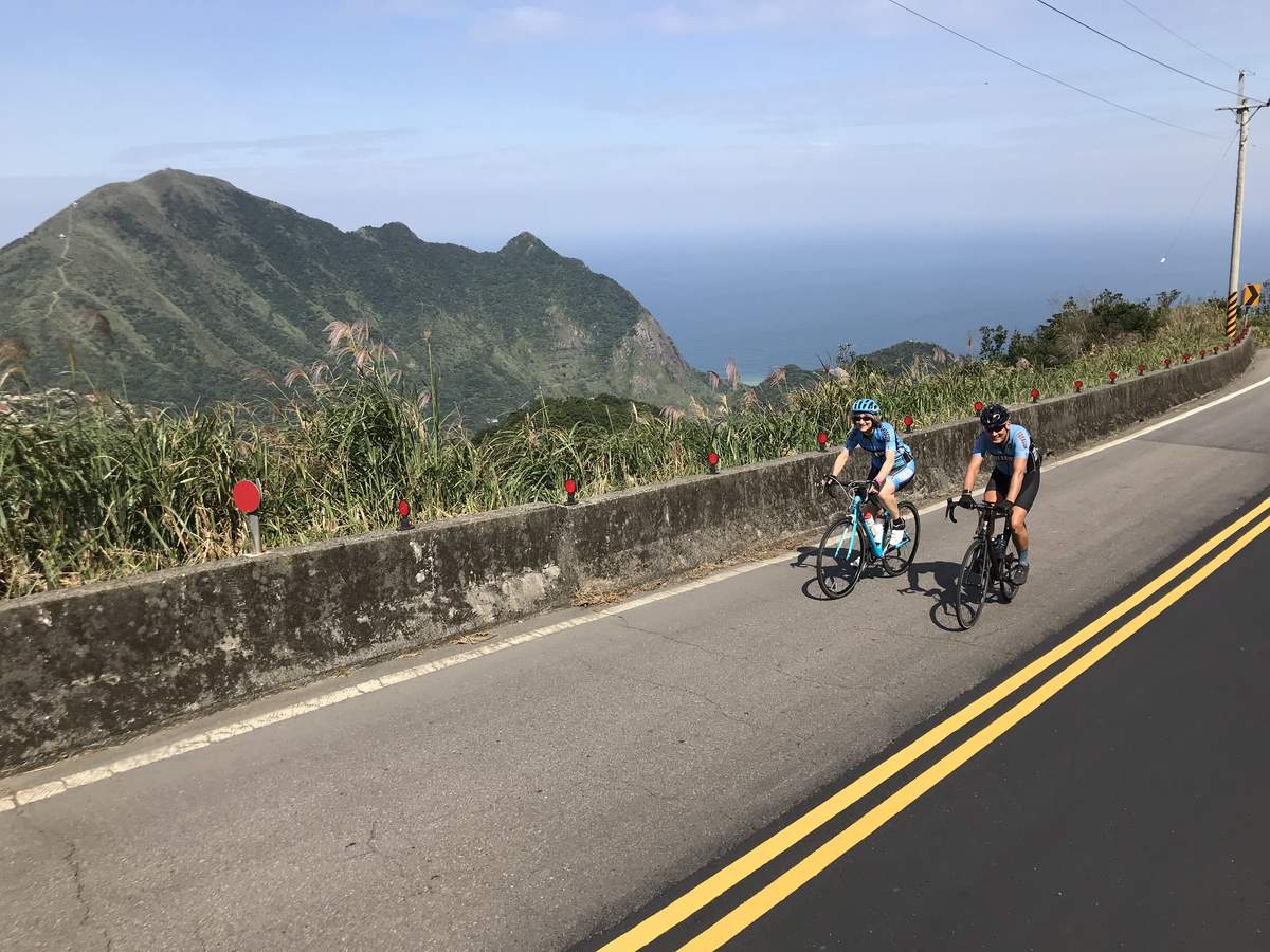 Two cyclists riding a coastal mountain road on the north-east coast of Taiwan, with a dramatic green headland and the Pacific Ocean in the background