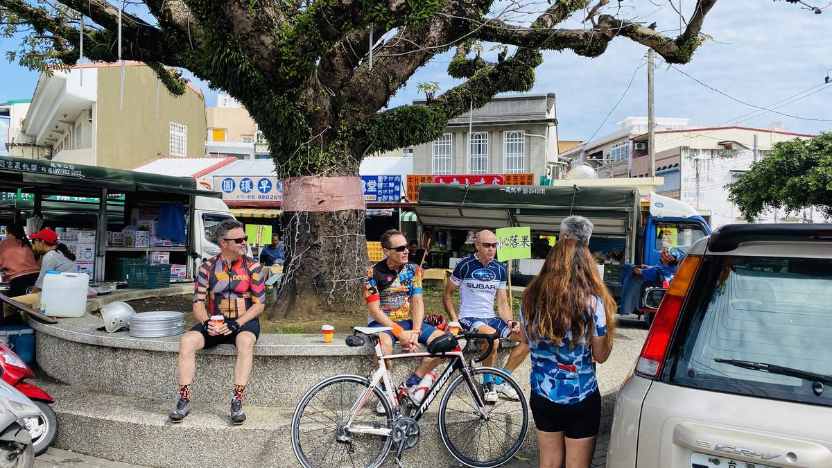 Three cyclists resting in the shade of a tree at a village square in Taiwan, with the support vehicle parked nearby and food stalls in the background