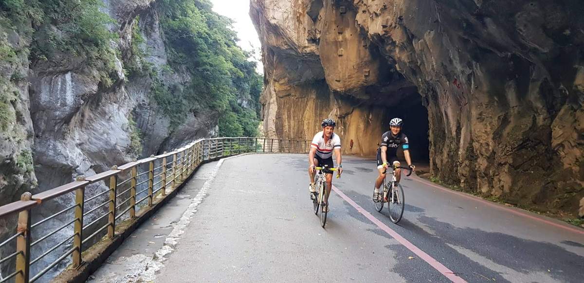 Two cyclists riding through the marble walls of Taroko Gorge with a stone tunnel cut into the cliff, on the lower Taiwan KOM route