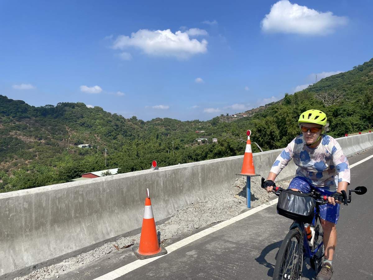 Solo cyclist on a flat-bar e-bike crossing a newly paved mountain road in Taiwan with forested hills behind and traffic cones marking the edge