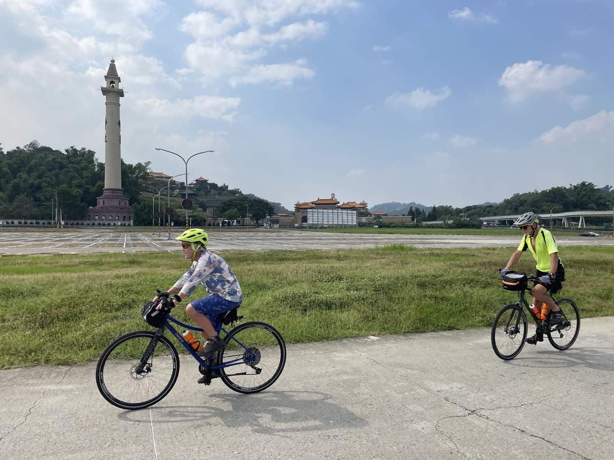 Two cyclists on flat-bar e-bikes riding a quiet path past a Taiwanese temple and pagoda across the river
