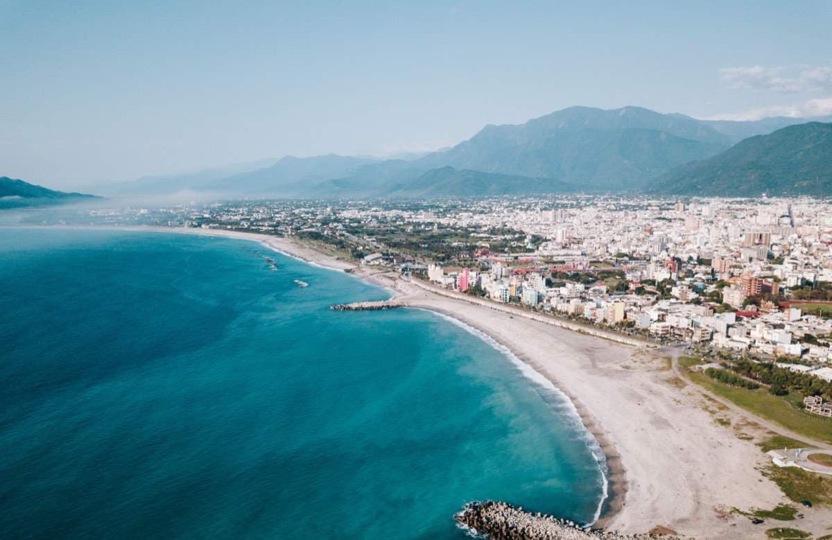 Aerial view of Taiwan's east coast, turquoise Pacific Ocean curving along the beach with the Coastal Mountain Range rising behind the town
