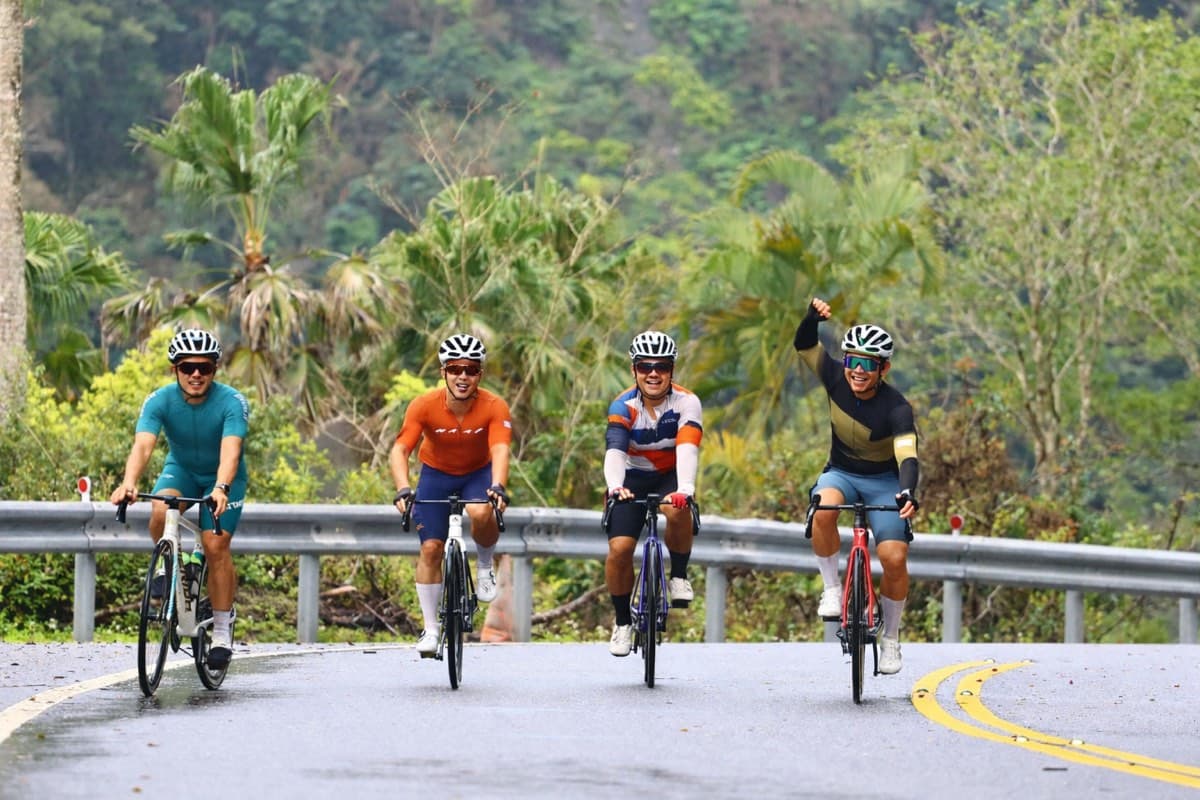 Group of four cyclists riding abreast and smiling on a tropical road in Taiwan, one rider with fist raised in celebration