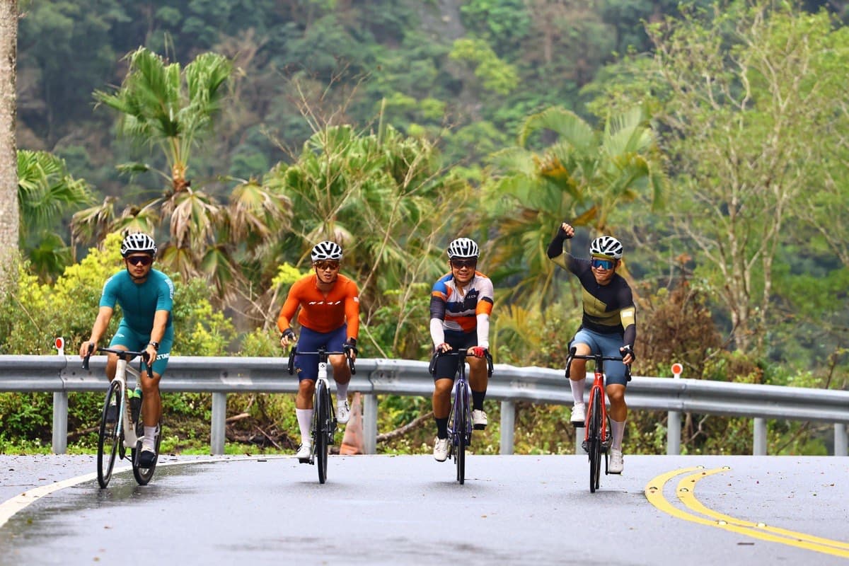 Four cyclists riding a sweeping tropical mountain road in Taiwan, wearing a mix of jerseys, bib shorts, helmets, and sunglasses