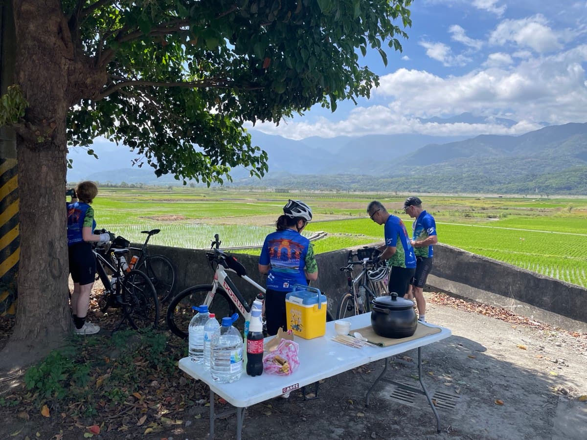 Pedal Taiwan group stopping for a BBQ lunch in the rift valley, bikes parked under a tree, rice paddies and mountains stretching out behind