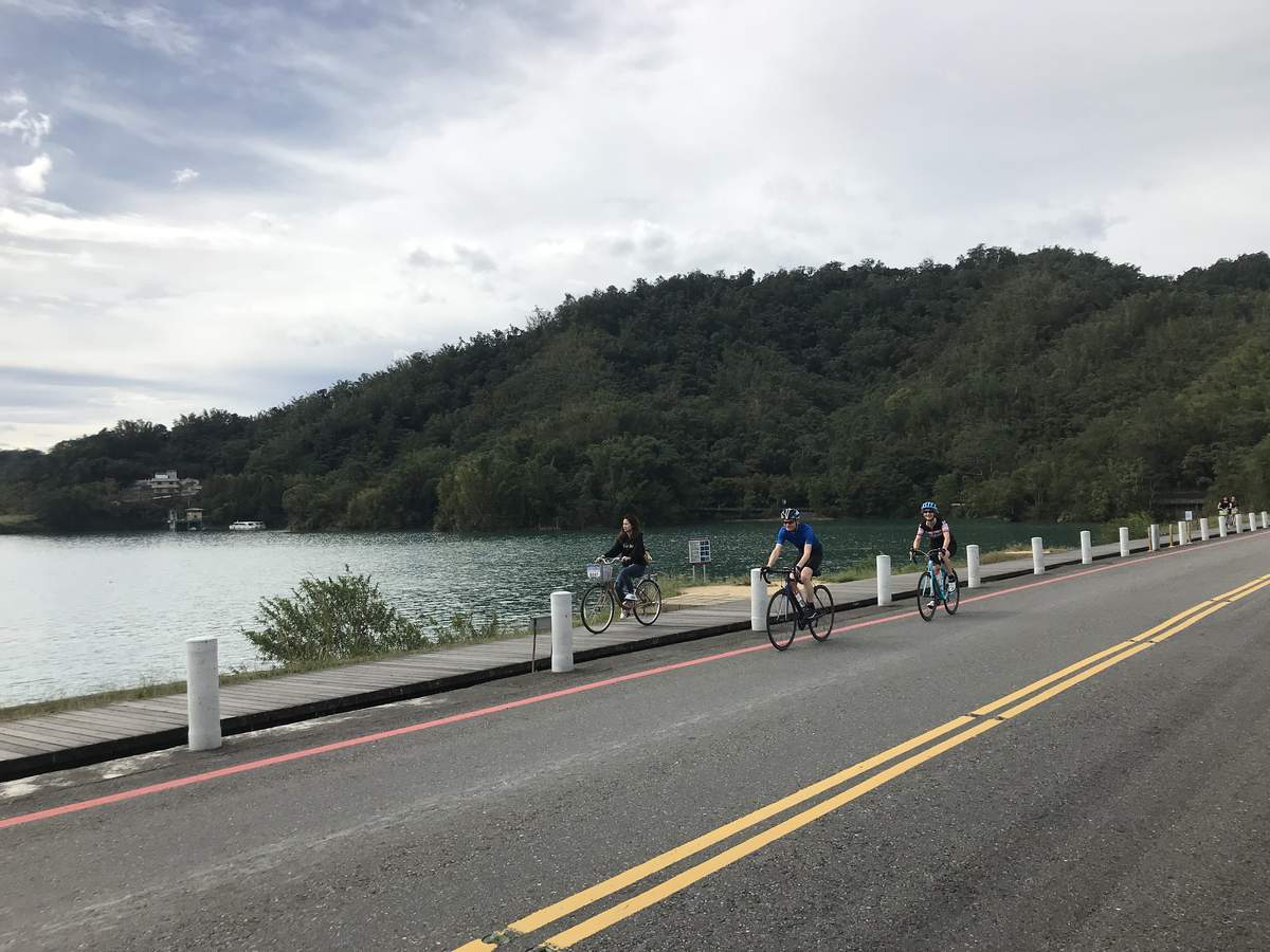Four cyclists riding on the dedicated Sun Moon Lake bike path beside the water with forested hills behind under a soft sky