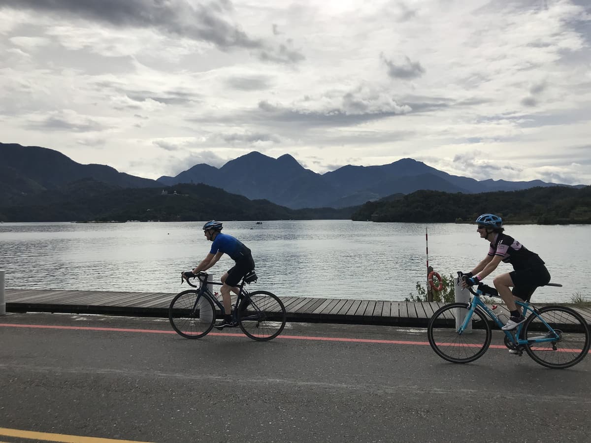 Two cyclists riding past Sun Moon Lake on the lakeside cycle path with the central mountain range and dramatic cloudy sky in the background
