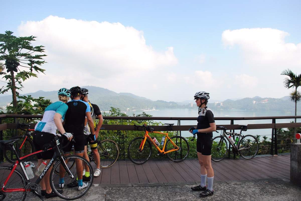 Group of cyclists with road bikes gathered at a scenic Sun Moon Lake overlook, the lake and forested mountains visible behind them under tropical sky